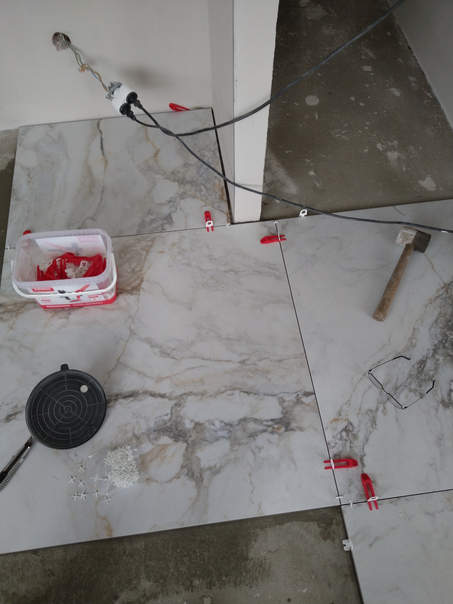 Układanie marmurowych płytek podłogowych z użyciem systemu poziomowania, widoczne narzędzia: młotek, przyssawka, krzyżyki dystansowe, poziomica, okulary i wiadro z klipsami.