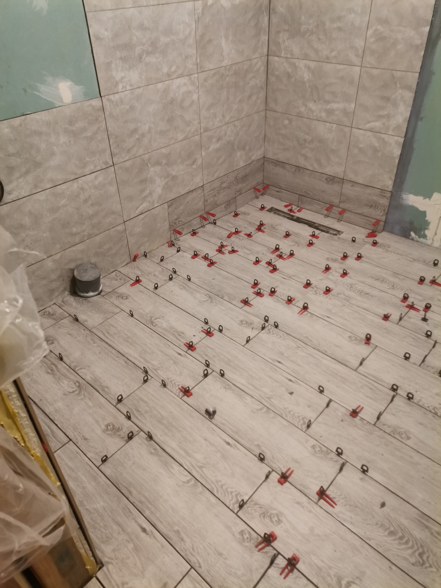 Układanie płytek imitujących drewno na podłodze w łazience w Szemudzie, z użyciem czerwonych klipsów poziomujących. Częściowo wykończone ściany z szarych, kwadratowych płytek.