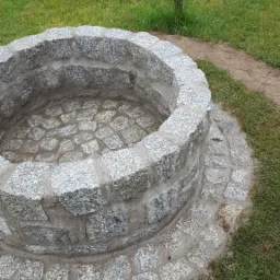 Okrągła studnia ogrodowa z jasnoszarej kostki granitowej, wykończona na poziomie gruntu, otoczona trawnikiem i młodym drzewkiem.