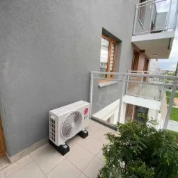 Biała jednostka zewnętrzna klimatyzatora marki LG zamontowana na czarnych podstawkach na balkonie apartamentu z widokiem na miasto.