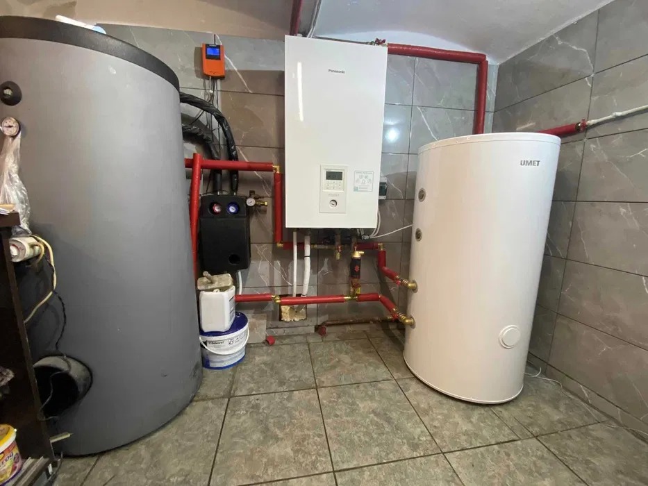 Instalacja kotła grzewczego Panasonic i dwóch zbiorników akumulacyjnych, połączonych rurami w kolorze czerwonym, na tle płytek imitujących marmur.