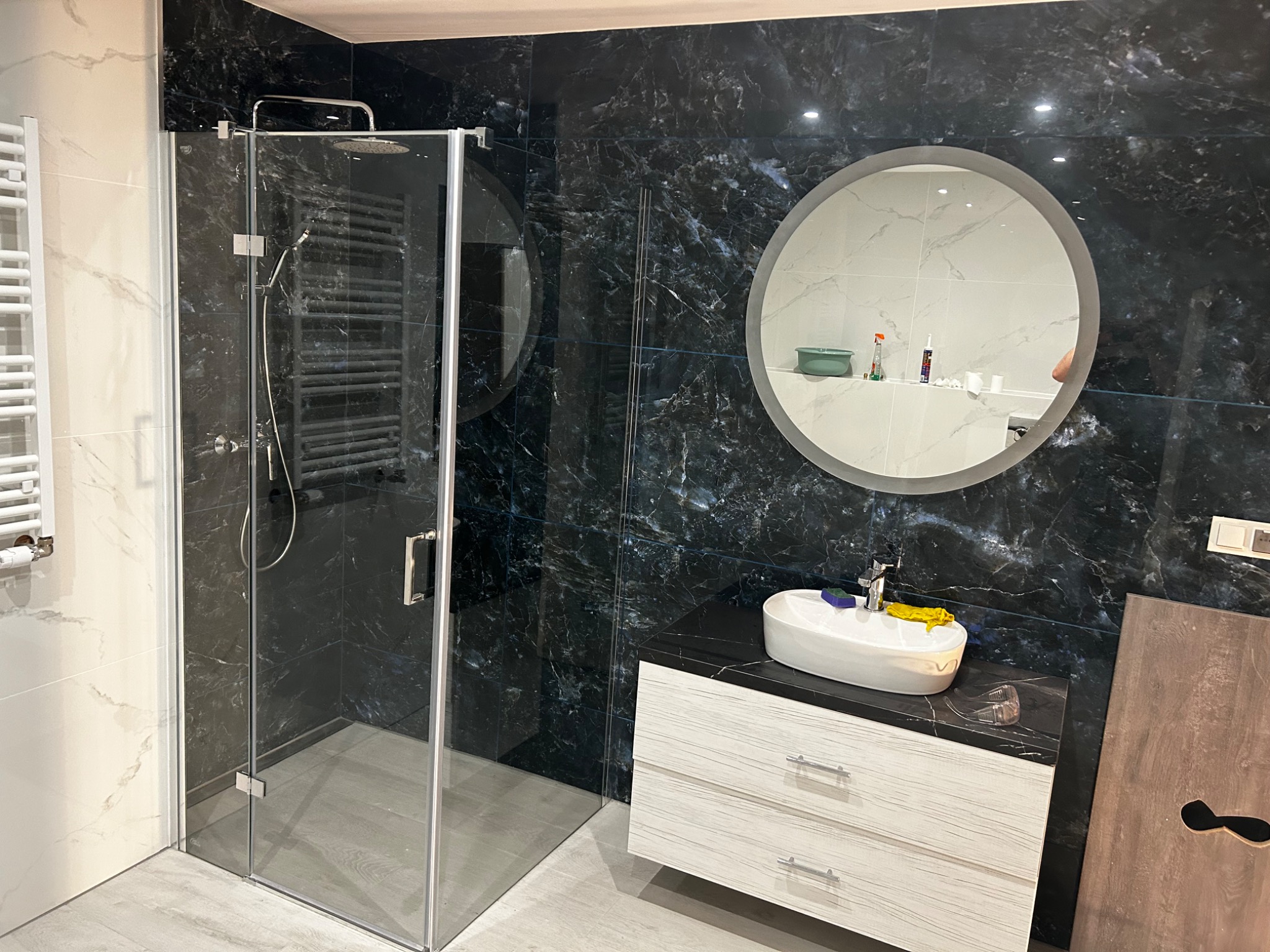 Nowoczesna łazienka z kabiną prysznicową ze szklanymi drzwiami, umywalką nablatową na szafce z szufladami i okrągłym lustrem na ścianie z czarnych płytek imitujących marmur.