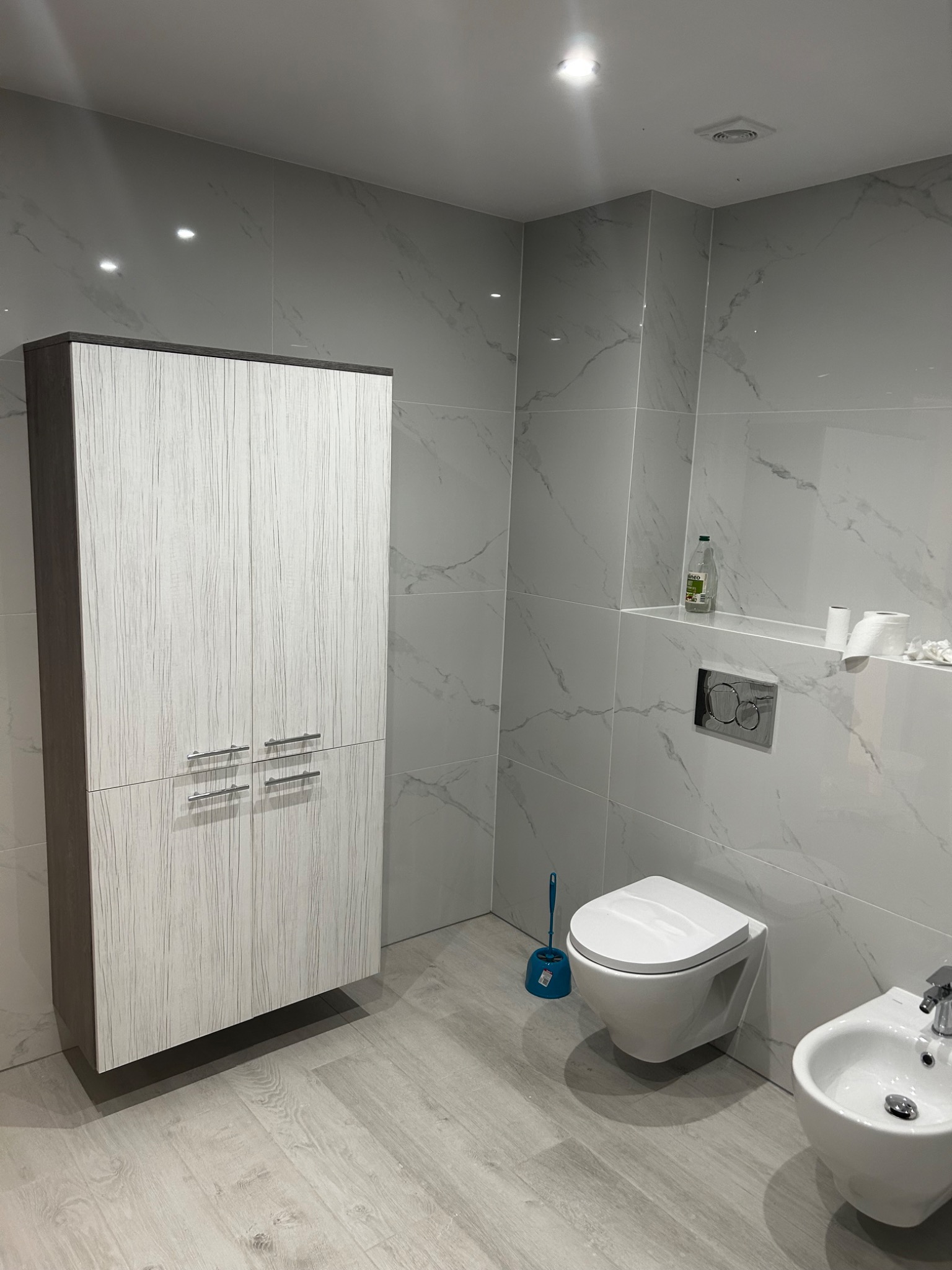 Nowoczesna łazienka z wiszącą toaletą i bidetem, szafką z drewnopodobnymi frontami, wykończona płytkami imitującymi marmur, z akcesoriami łazienkowymi.