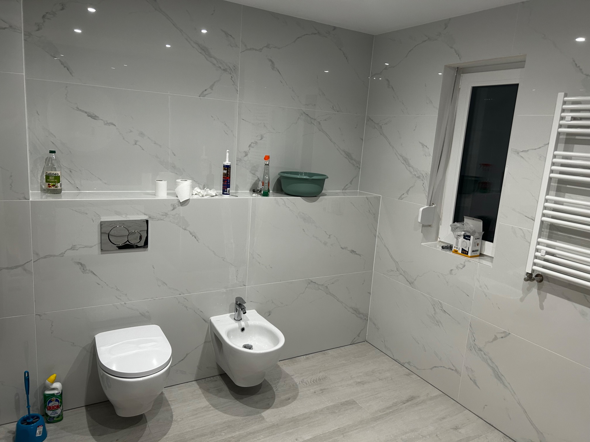 Wykończona łazienka w Kostrzynie z białymi płytkami imitującymi marmur, wiszącą toaletą, bidetem i grzejnikiem drabinkowym. Na półce widoczne środki czystości i akcesoria.