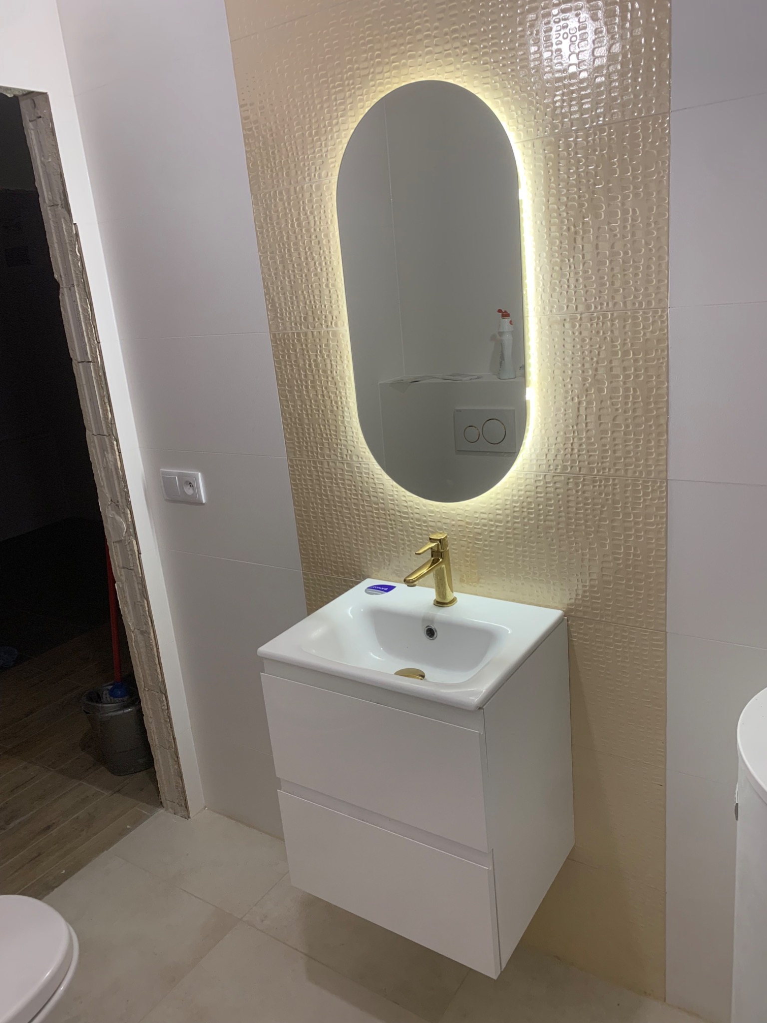 Nowoczesna łazienka z umywalką na szafce, złotą baterią i podświetlanym lustrem owalnym, widoczne płytki strukturalne w odcieniach beżu i białe ściany, fragment niedokończonego wejścia.