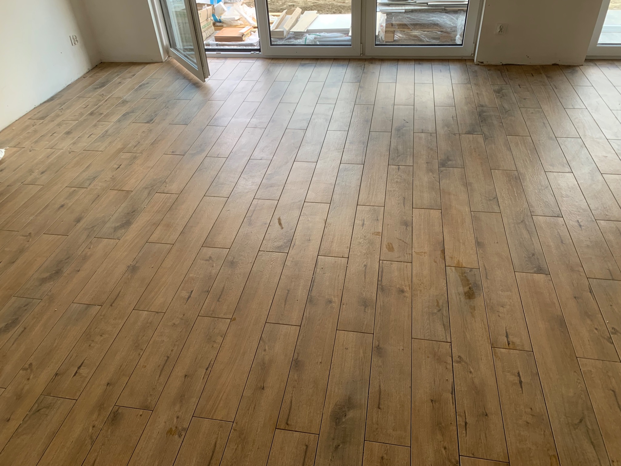 Ułożona podłoga z płytek imitujących drewno w nowym mieszkaniu, widok na rozsuwane drzwi balkonowe i materiały budowlane w tle.