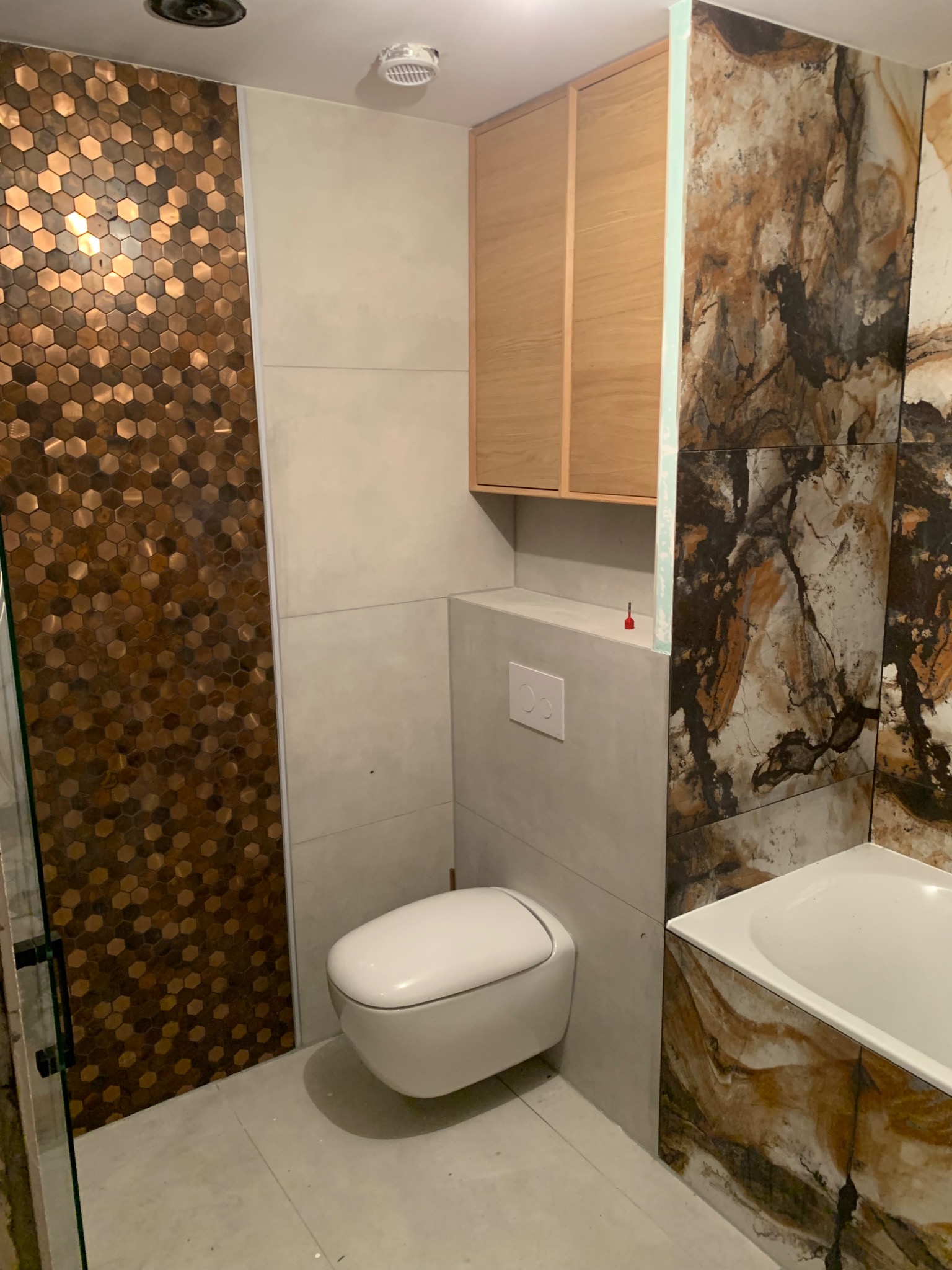 Wnętrze łazienki w trakcie wykończenia: mozaika heksagonalna, płytki imitujące marmur, podwieszana toaleta, zabudowa geberit, szafka wisząca z jasnego drewna.