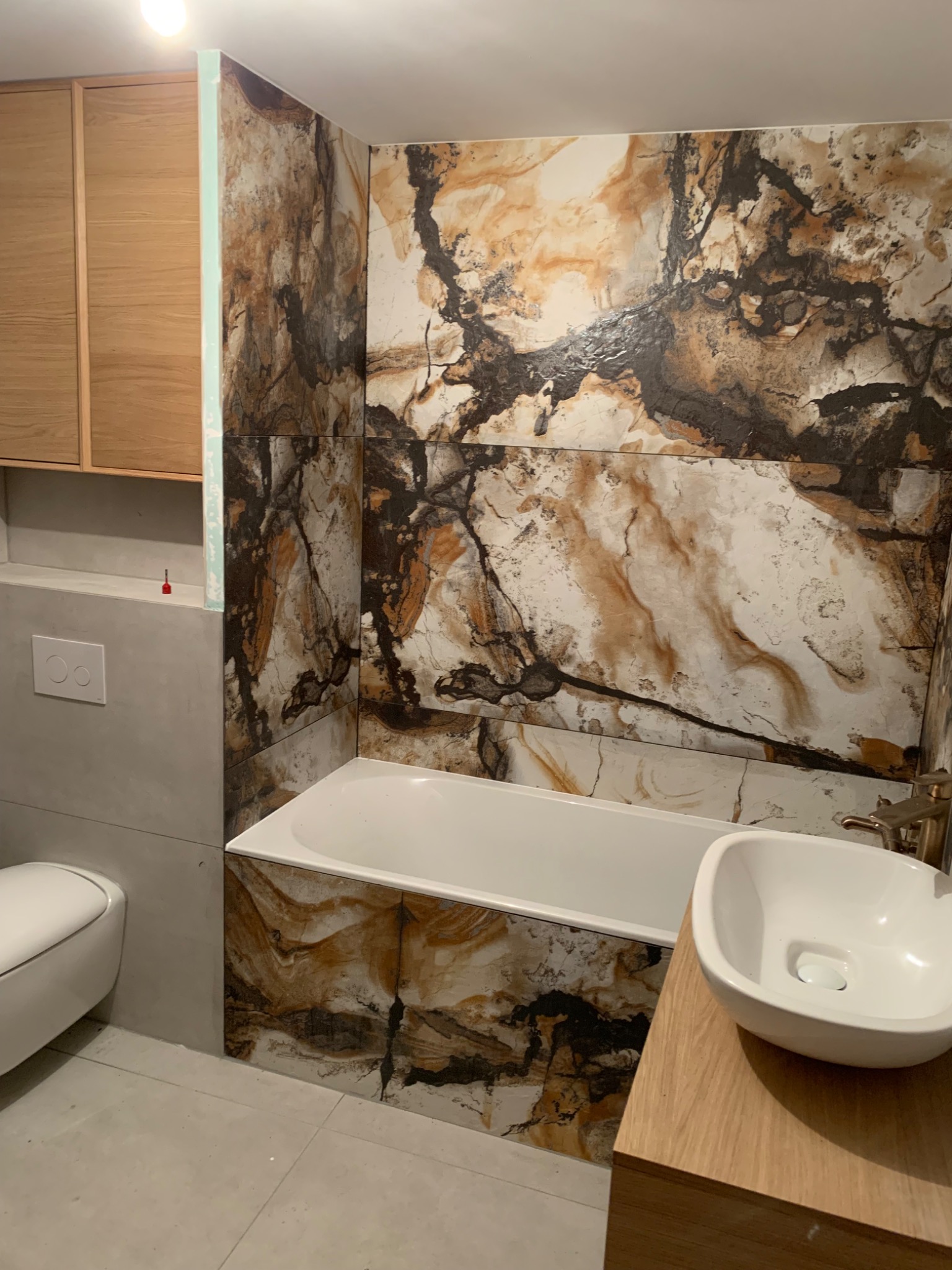 Nowoczesna łazienka z marmurowymi płytkami, białą wanną, umywalką nablatową na drewnianej szafce i podwieszaną toaletą. Ciemne fugi podkreślają wzór płytek.