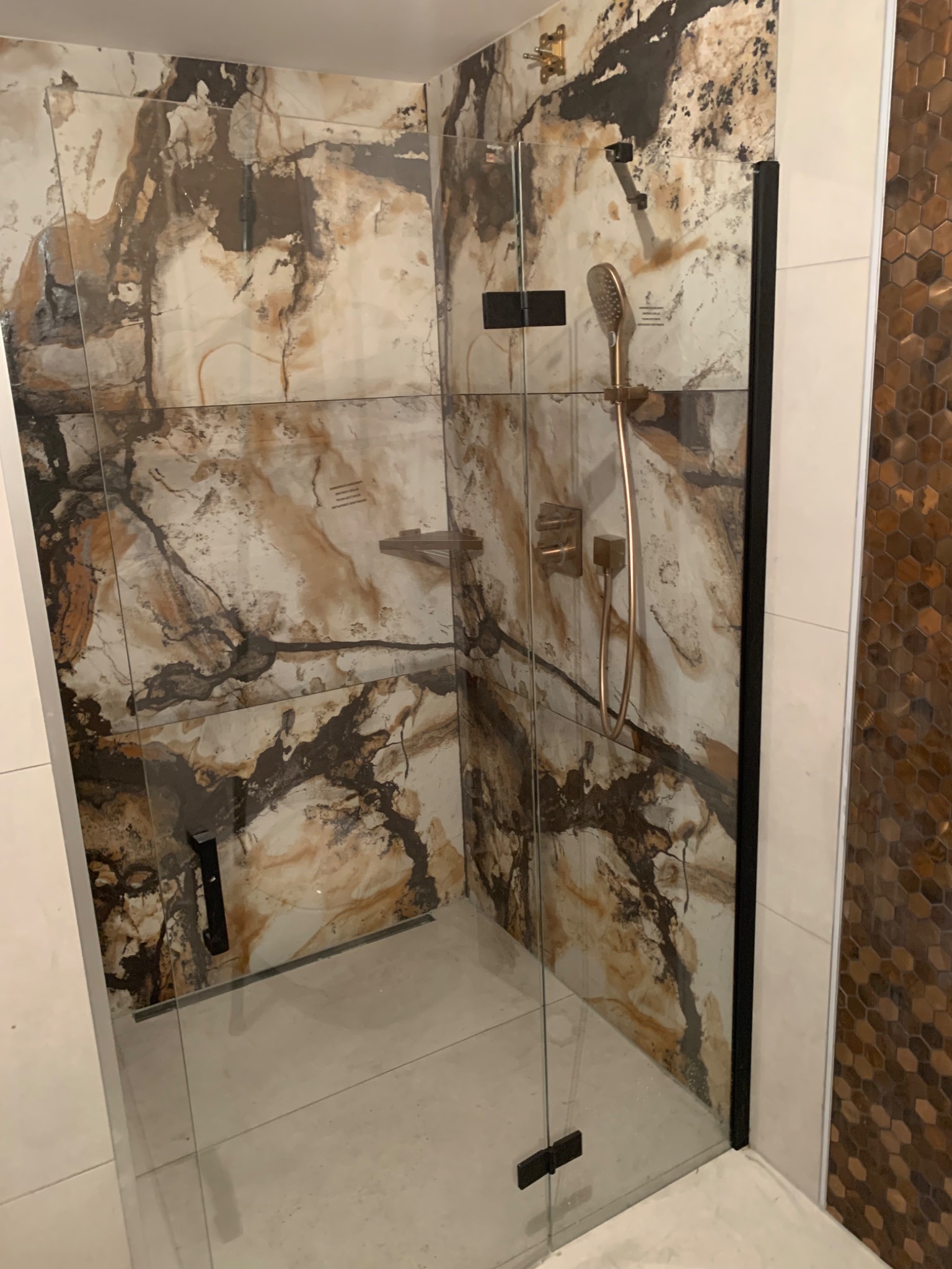 Kabina prysznicowa walk-in z czarnymi profilami i szklanymi drzwiami, marmurowe płytki na ścianach, armatura w kolorze mosiądzu, heksagonalna mozaika na sąsiedniej ścianie.