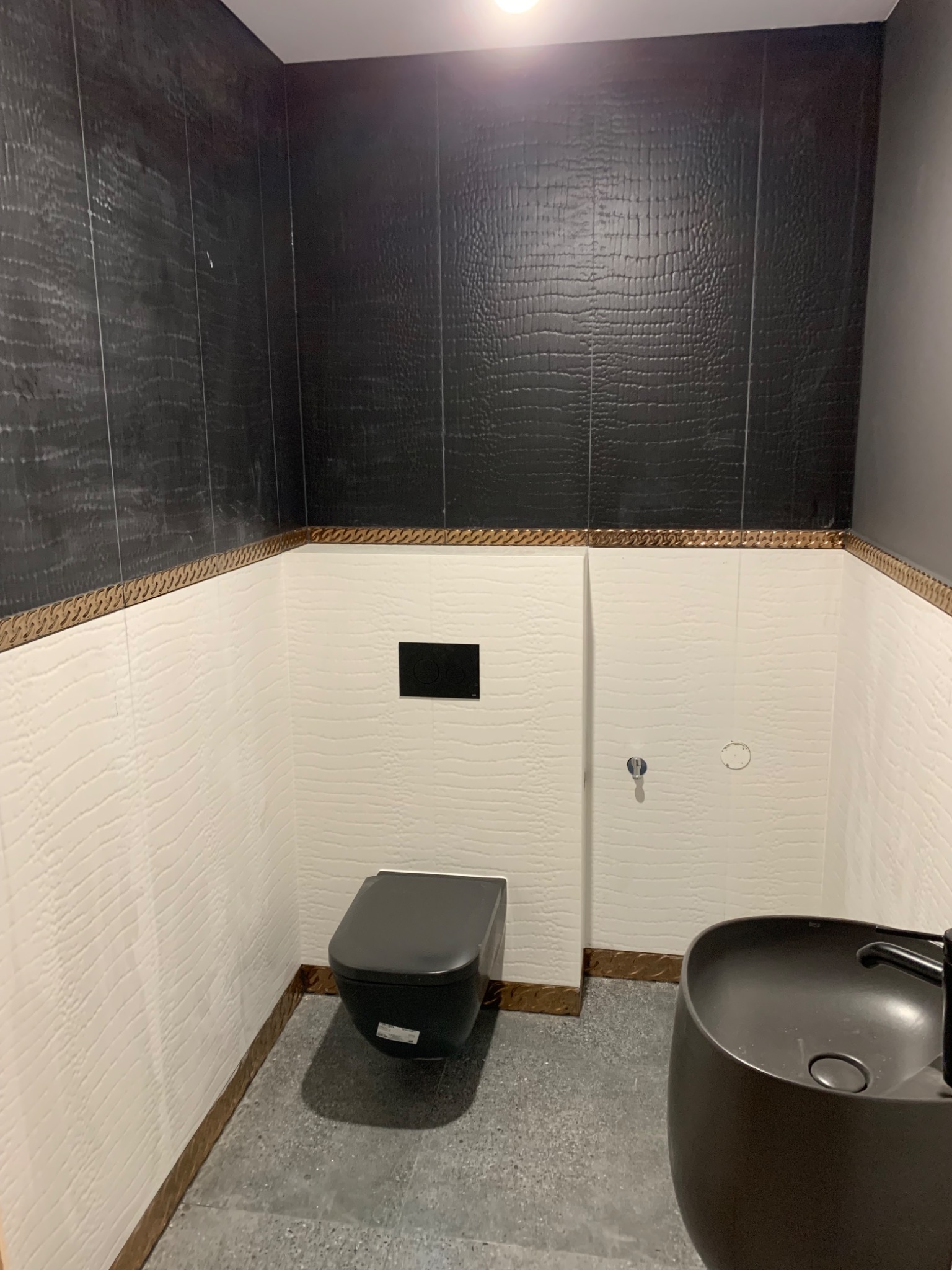 Nowoczesna łazienka z czarnymi płytkami imitującymi skórę krokodyla na górnej części ścian i białymi na dole, oddzielonymi ozdobnym pasem, z czarną umywalką i toaletą.