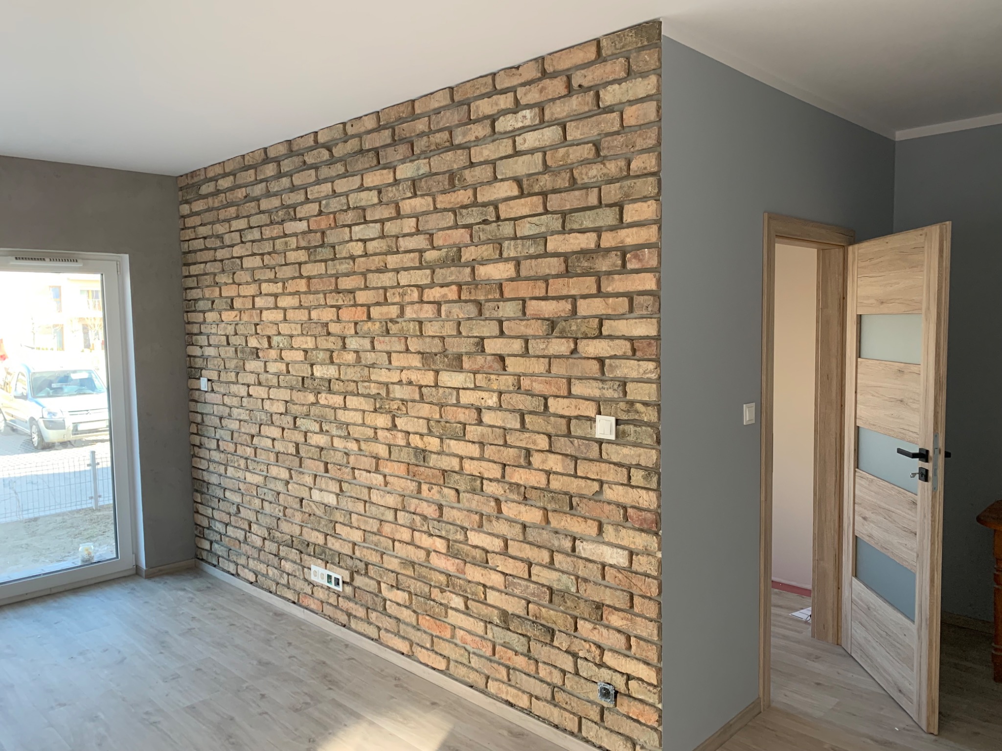 Ściana z cegły dekoracyjnej w nowocześnie wykończonym wnętrzu, szara ściana boczna, jasna podłoga, otwarte drzwi wewnętrzne z przeszkleniami.