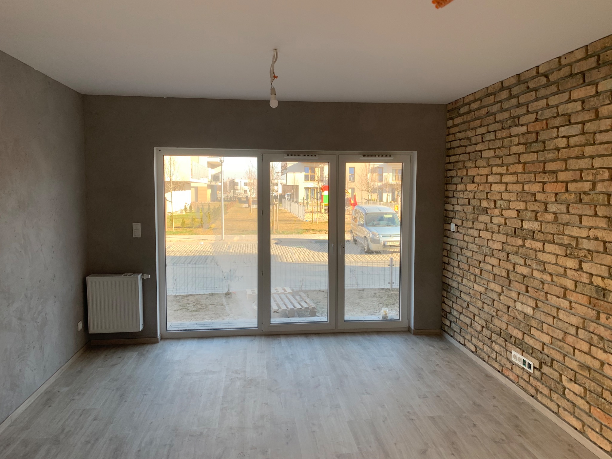 Pusty pokój po remoncie z panelami podłogowymi, ścianą z cegły dekoracyjnej, ścianą wykończoną gładzią i dużym oknem balkonowym.