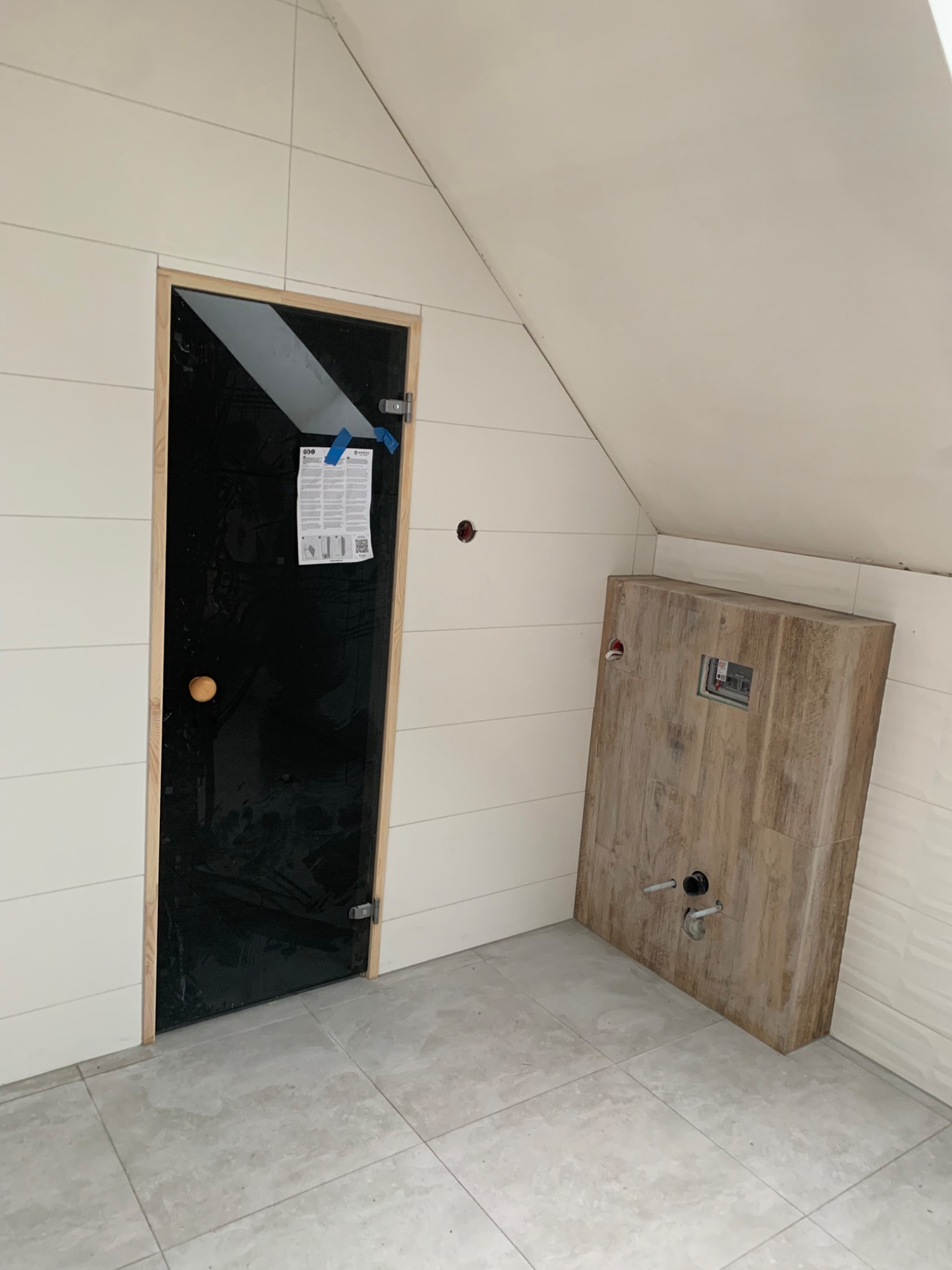 Łazienka na poddaszu w trakcie prac: zabudowa stelaża WC z imitacji drewna, białe płytki imitujące panele na ścianach, czarne drzwi z drewnianą ościeżnicą zabezpieczone folią.