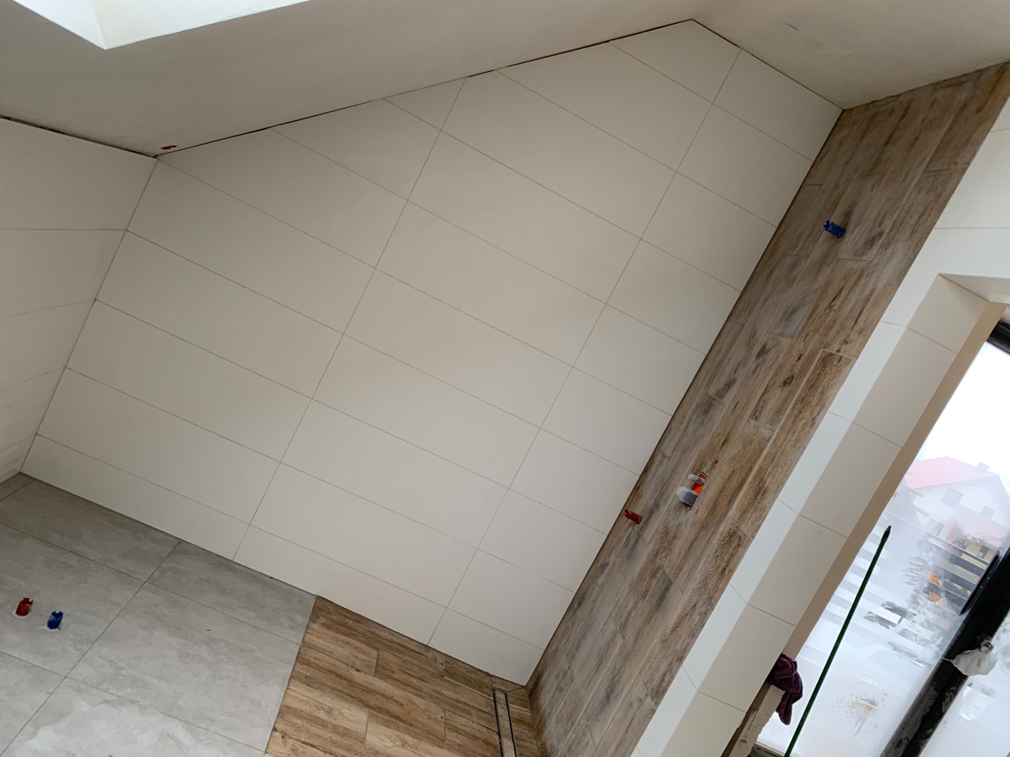 Ukończona łazienka na poddaszu z białymi płytkami na skośnej ścianie i drewnopodobnymi płytkami na fragmencie ściany oraz podłodze, z widocznymi elementami mocującymi płytki.