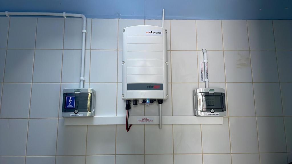 Instalacja fotowoltaiczna w pomieszczeniu: centralny inwerter NexEnergy Solar i dwa panele sterujące na białej, kafelkowanej ścianie, z widocznymi przewodami i ostrzeżeniem o podwójnym zasilaniu.