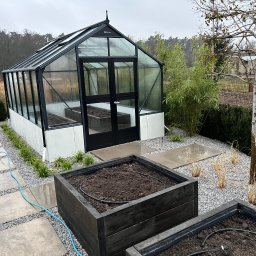 Usługi Ogrodnicze Toruń - Ogrody Nowiccy F.H.U Tatiana Walkiewicz Nowicka - Szklarnia z czarną ramą i podwyższone grządki z ciemnego drewna, wyposażone w system nawadniający, otoczone szarym żwirem i płytami chodnikowymi.