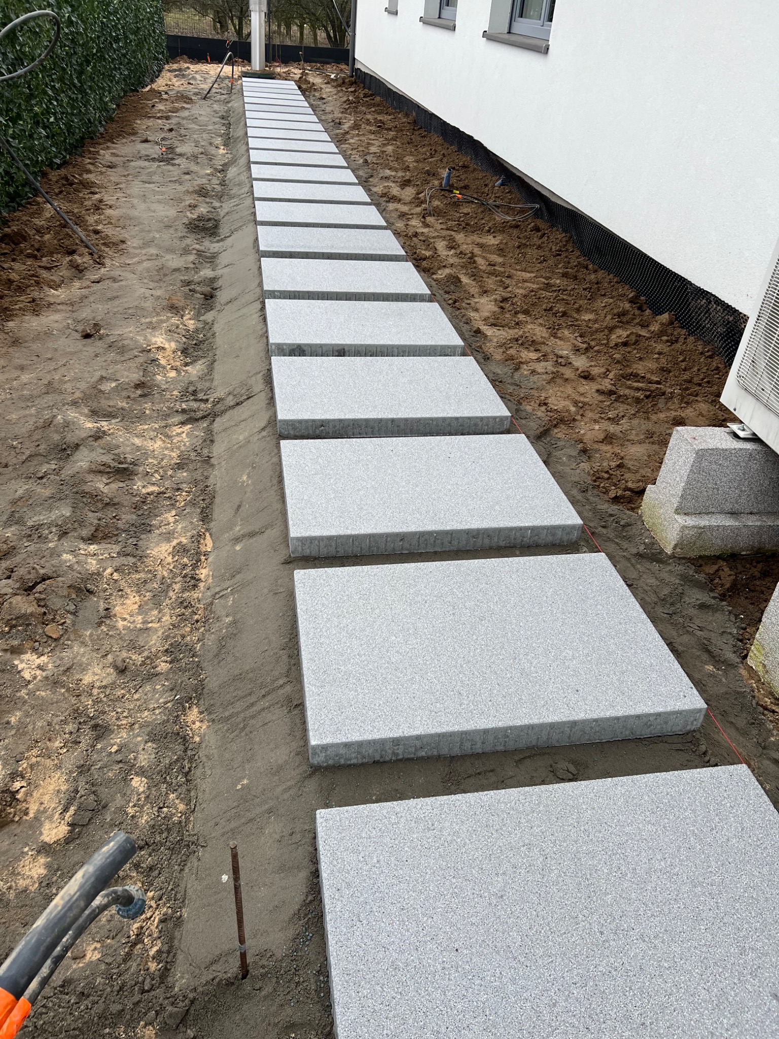 Ścieżka z szarych, kwadratowych płyt ułożona wzdłuż ściany budynku, widoczne przygotowane podłoże i narzędzia ogrodnicze.