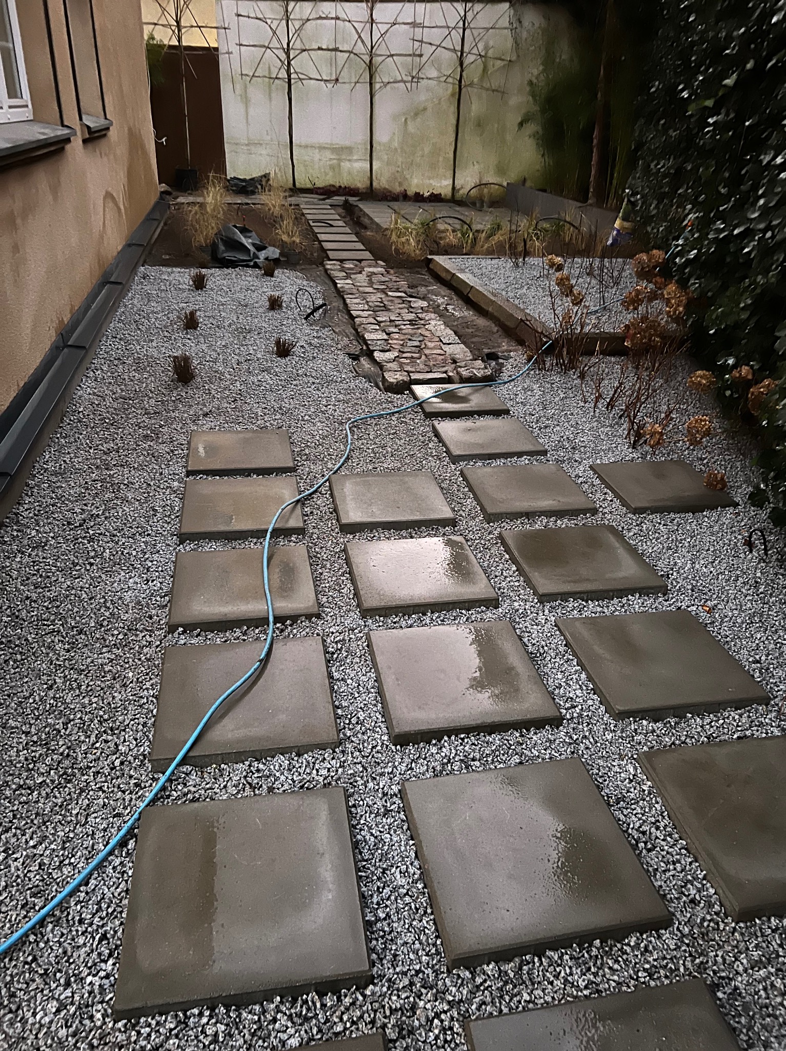 Ścieżka z kwadratowych płyt ułożonych na szarym żwirze w ogrodzie, z widocznym niebieskim wężem ogrodowym i fragmentem brukowanej ścieżki w tle.