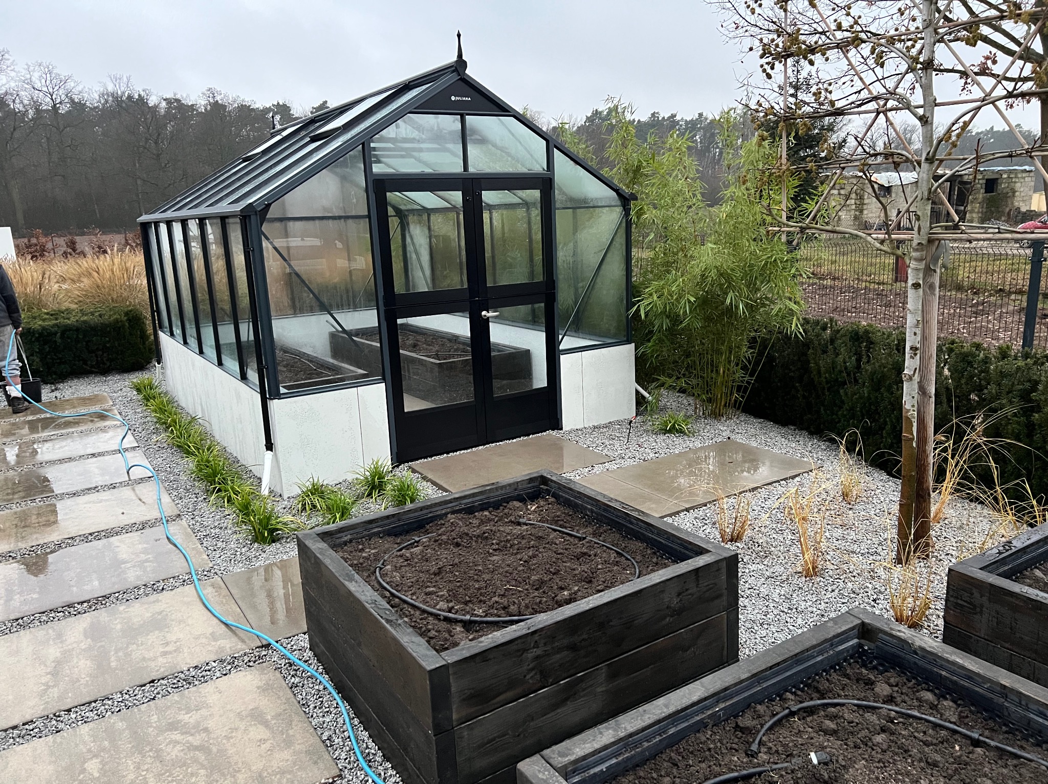 Szklarnia z czarną ramą i podwyższone grządki z ciemnego drewna, wyposażone w system nawadniający, otoczone szarym żwirem i płytami chodnikowymi.