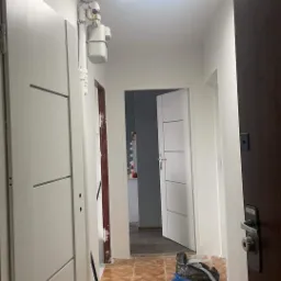 Korytarz w trakcie prac wykończeniowych: białe ściany, nowe drzwi z frezowanymi panelami, nowoczesna lampa LED, widoczny licznik gazu, brązowa podłoga z terakoty, narzędzia do sprzątania i worek...