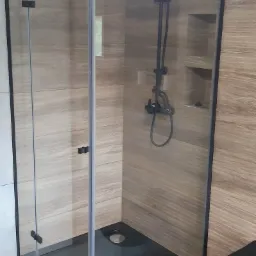 Nowoczesna kabina prysznicowa z czarnym brodzikiem i armaturą, wykończona płytkami imitującymi drewno, z widoczną częścią toalety w tle.