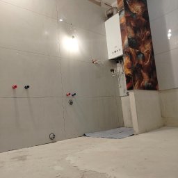 MARCIN GENEROWICZ USŁUGI REMONTOWO-WYKOŃCZENIOWE - Łazienka w trakcie wykończenia: szare płytki na ścianach, dekoracyjny panel z efektem marmuru obok pieca gazowego. Widoczne przyłącza wodne i surowa podłoga.