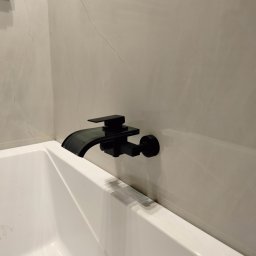 MARCIN GENEROWICZ USŁUGI REMONTOWO-WYKOŃCZENIOWE - Bateria wannowa w kolorze czarnym, zamontowana na ścianie z jasnych, marmurkowych płytek. Widoczny fragment białej wanny. Minimalistyczny design.