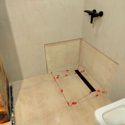 MARCIN GENEROWICZ USŁUGI REMONTOWO-WYKOŃCZENIOWE - Montaż odpływu liniowego w łazience z beżowymi płytkami imitującymi marmur. Widoczne krzyżyki glazurnicze i czarna bateria. Fragment umywalki w kadrze.