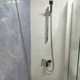 Wykończona kabina prysznicowa z szarymi płytkami imitującymi marmur i jasnymi płytkami. Widoczna chromowana armatura i szklane drzwi kabiny.