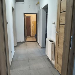 MARCIN GENEROWICZ USŁUGI REMONTOWO-WYKOŃCZENIOWE - Wykończony korytarz z szarą, kwadratową podłogą, białymi ścianami i drewnianymi drzwiami. Widoczny grzejnik i fragment pomieszczenia w trakcie remontu w tle.