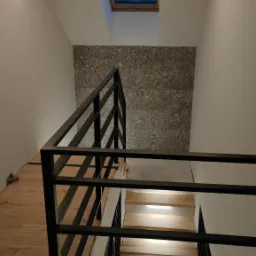 Schody drewniane z czarną metalową balustradą, oświetlone od spodu, prowadzące na poddasze z oknem dachowym i ścianą wykończoną płytkami imitującymi lastryko.