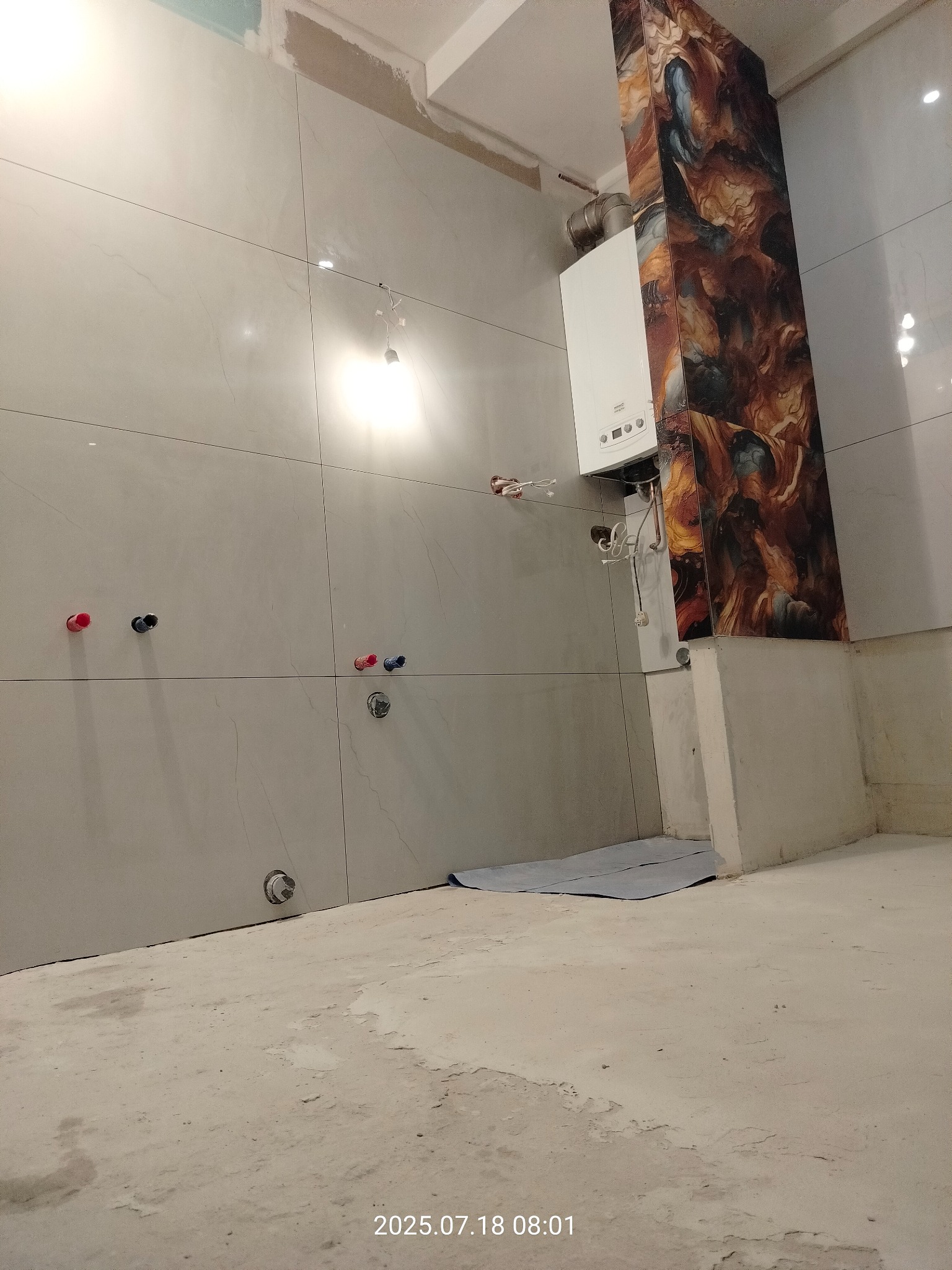 Łazienka w trakcie wykończenia: szare płytki na ścianach, dekoracyjny panel z efektem marmuru obok pieca gazowego. Widoczne przyłącza wodne i surowa podłoga.