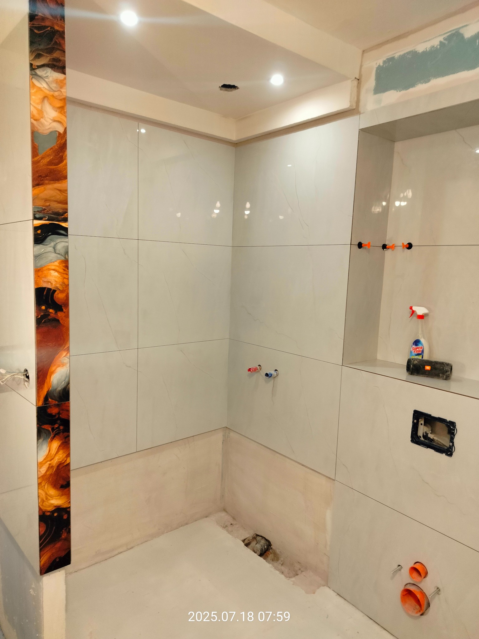 Wnętrze łazienki w trakcie prac wykończeniowych: białe płytki imitujące marmur, dekoracyjny panel z żywicy epoksydowej, widoczne elementy instalacji.