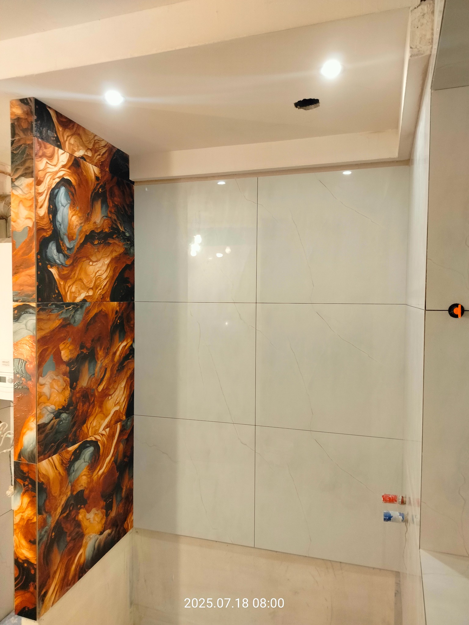 Wykończona ściana łazienki z białych, kwadratowych płytek i fragmentu ściany z płytkami imitującymi wzór marmuru. Widoczne elementy instalacji wodnej.