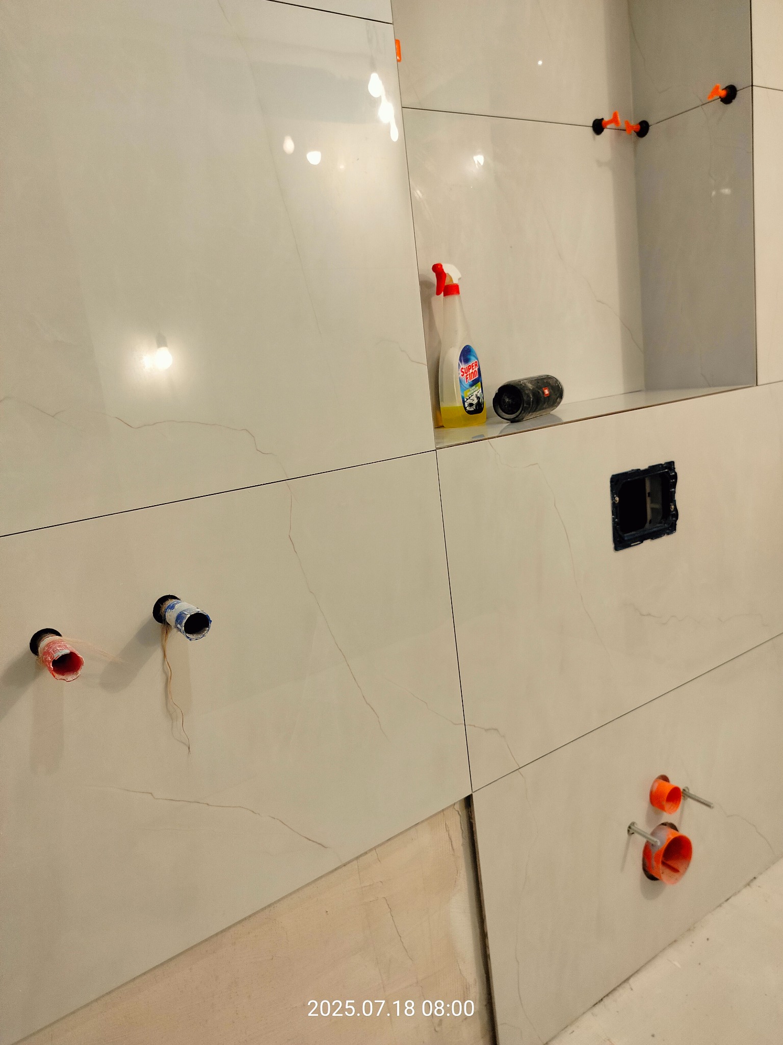 Ściana łazienki w trakcie wykańczania: białe płytki imitujące marmur, widoczne rury, puszka elektryczna, poziomica, środek czyszczący i system poziomowania płytek.