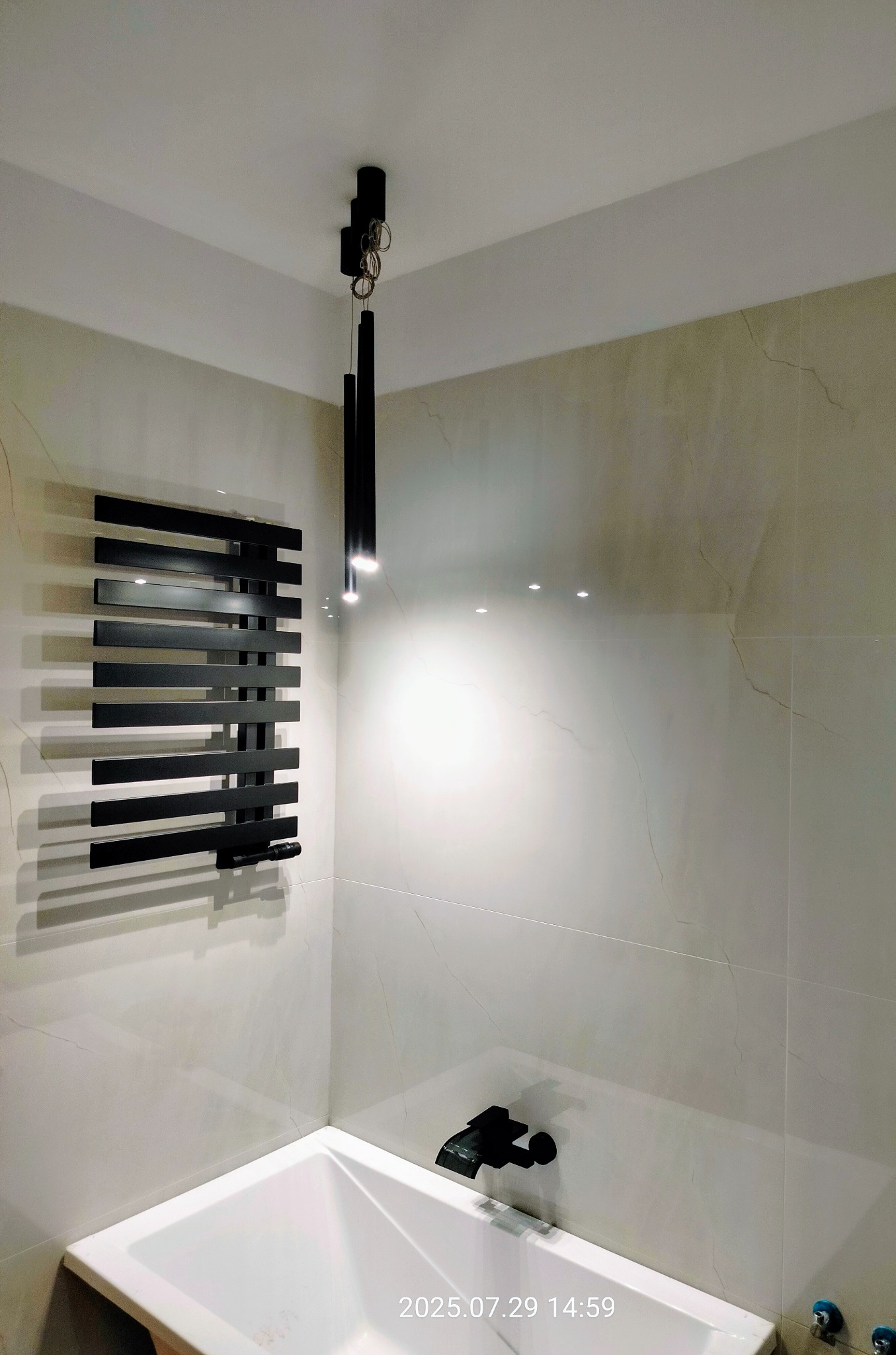 Nowoczesna łazienka z beżowymi płytkami imitującymi marmur, czarnym grzejnikiem drabinkowym i minimalistyczną lampą wiszącą. Ujęcie na wannę i armaturę.