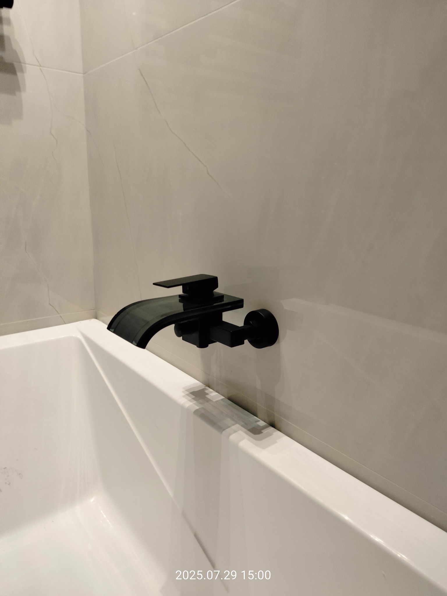 Bateria wannowa w kolorze czarnym, zamontowana na ścianie z jasnych, marmurkowych płytek. Widoczny fragment białej wanny. Minimalistyczny design.