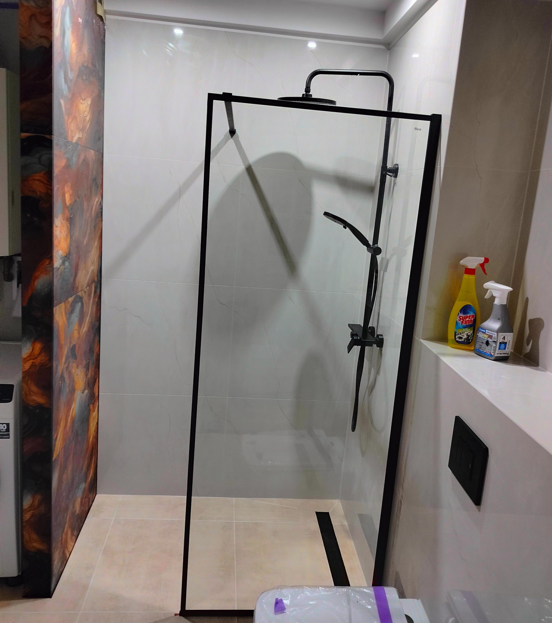 Nowoczesna kabina prysznicowa walk-in z czarnymi profilami, na tle jasnych płytek. Widoczny panel dekoracyjny z efektem marmuru i czarna armatura.