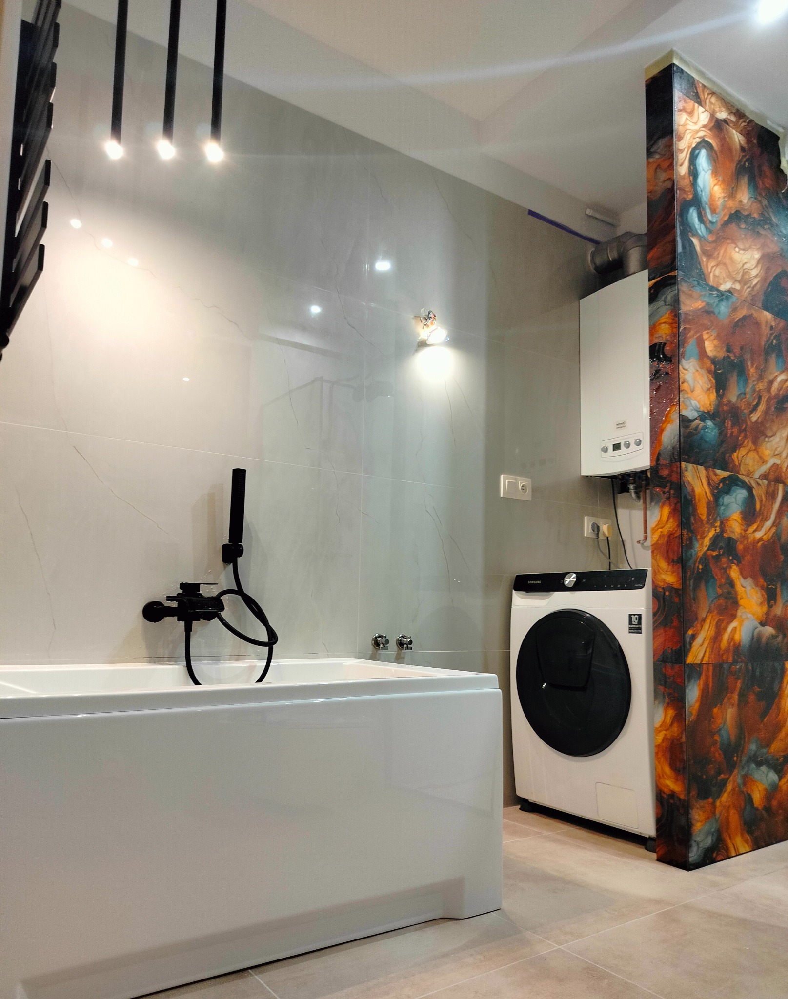 Nowoczesna łazienka z wanną, czarną armaturą i pralką. Marmurowe płytki na ścianach, dekoracyjny panel z efektem marmuru. Oświetlenie punktowe.