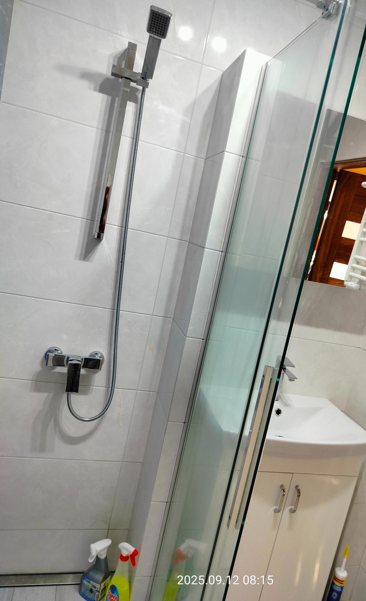 Nowoczesna kabina prysznicowa z białymi płytkami i chromowaną armaturą. Widoczne środki czystości. Ujęcie z dołu, eksponujące detale wykończenia.