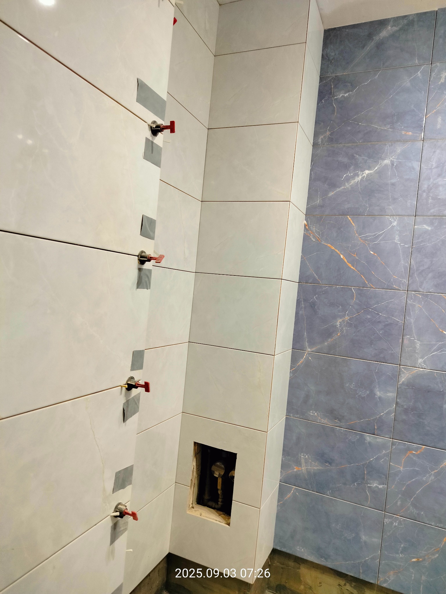 Układanie białych i grafitowych płytek imitujących marmur w łazience, z użyciem systemu poziomowania płytek z czerwonymi klinami i szarą taśmą maskującą.