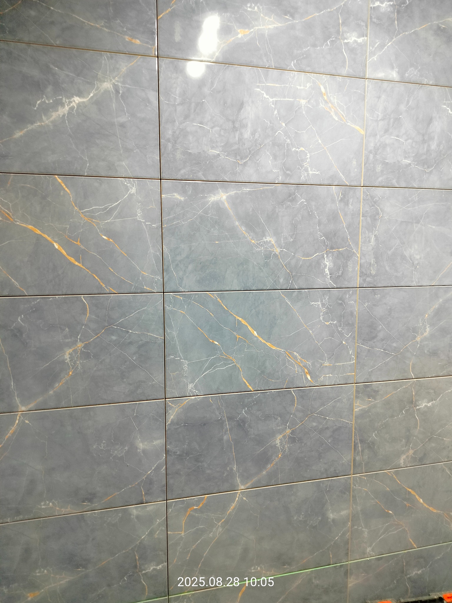 Szara ściana wyłożona prostokątnymi płytkami imitującymi marmur z delikatnymi złotymi żyłkami. Widoczne fugi podkreślają regularny układ.