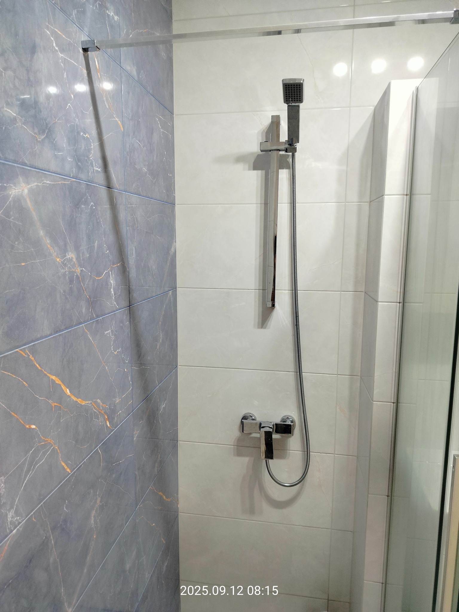 Wykończona kabina prysznicowa z szarymi płytkami imitującymi marmur i jasnymi płytkami. Widoczna chromowana armatura i szklane drzwi kabiny.