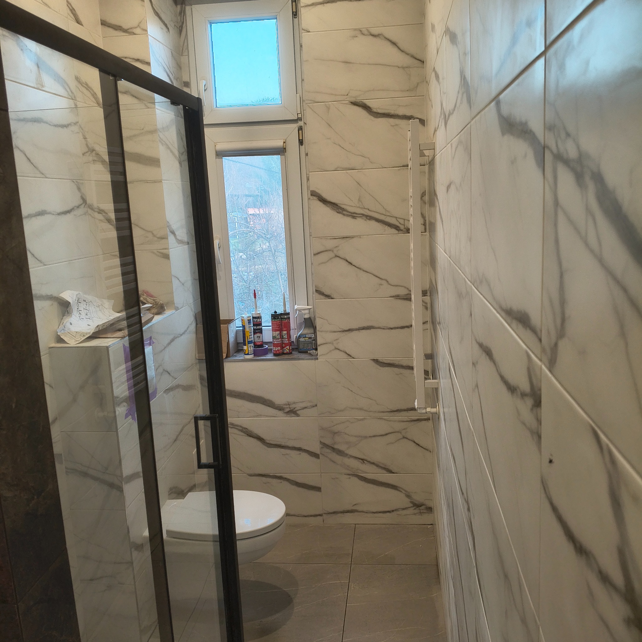 Wykończona łazienka z kabiną prysznicową typu walk-in, białe płytki imitujące marmur na ścianach, szara podłoga i okno z widokiem na zewnątrz.