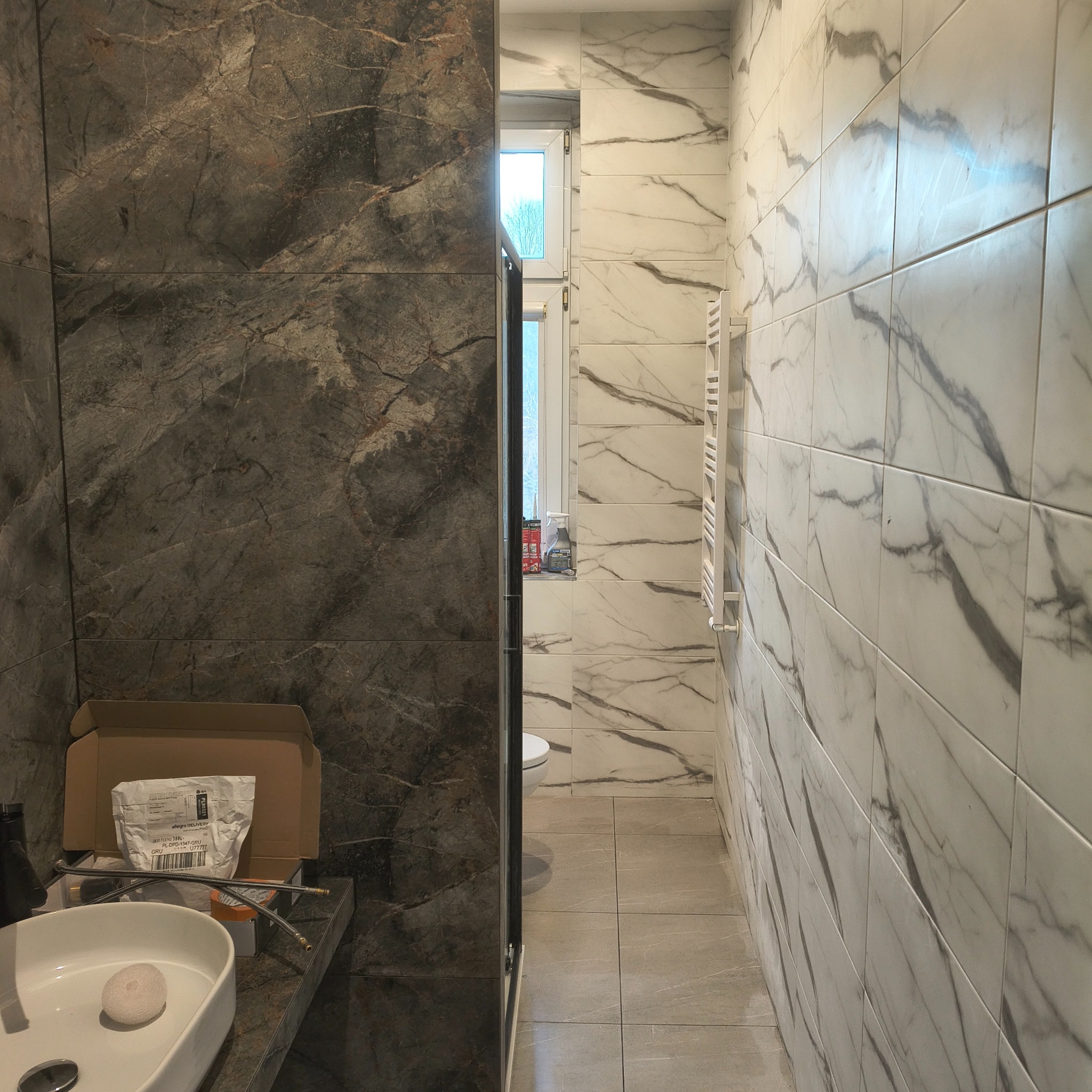 Wykończona łazienka z marmurowymi płytkami w odcieniach szarości i bieli. Widoczna umywalka, grzejnik i okno. Częściowo widoczne akcesoria montażowe.