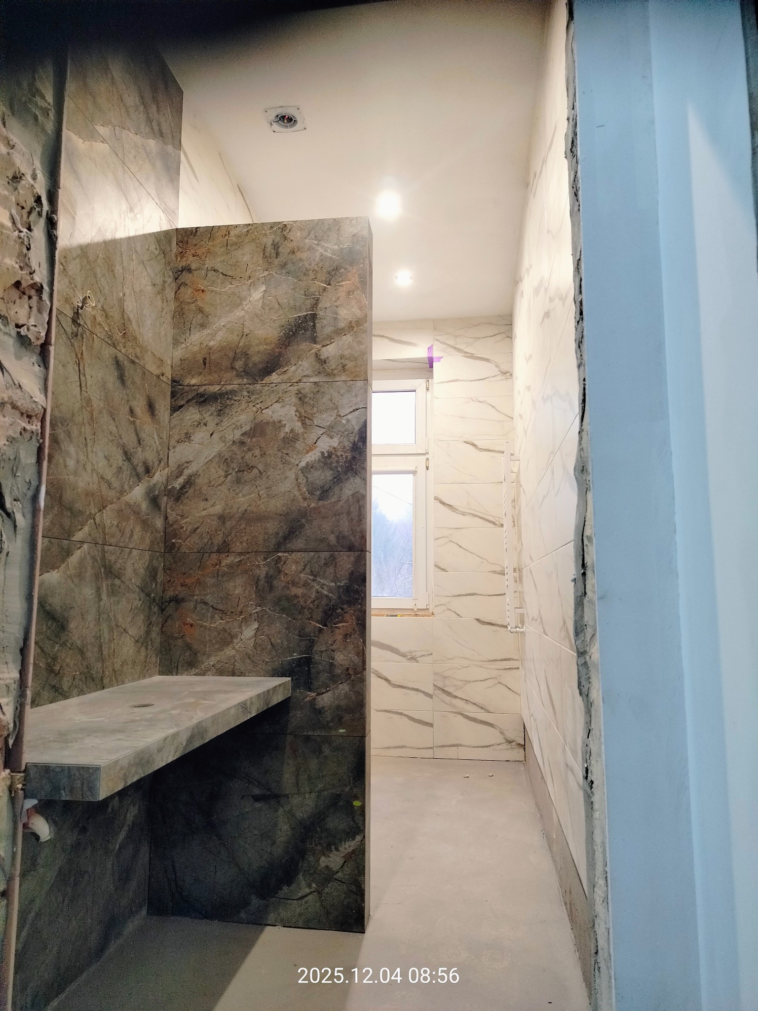 Wykończona łazienka z marmurowymi płytkami, widoczny fragment kabiny prysznicowej z półką i okno. Ściany wyłożone płytkami imitującymi marmur.