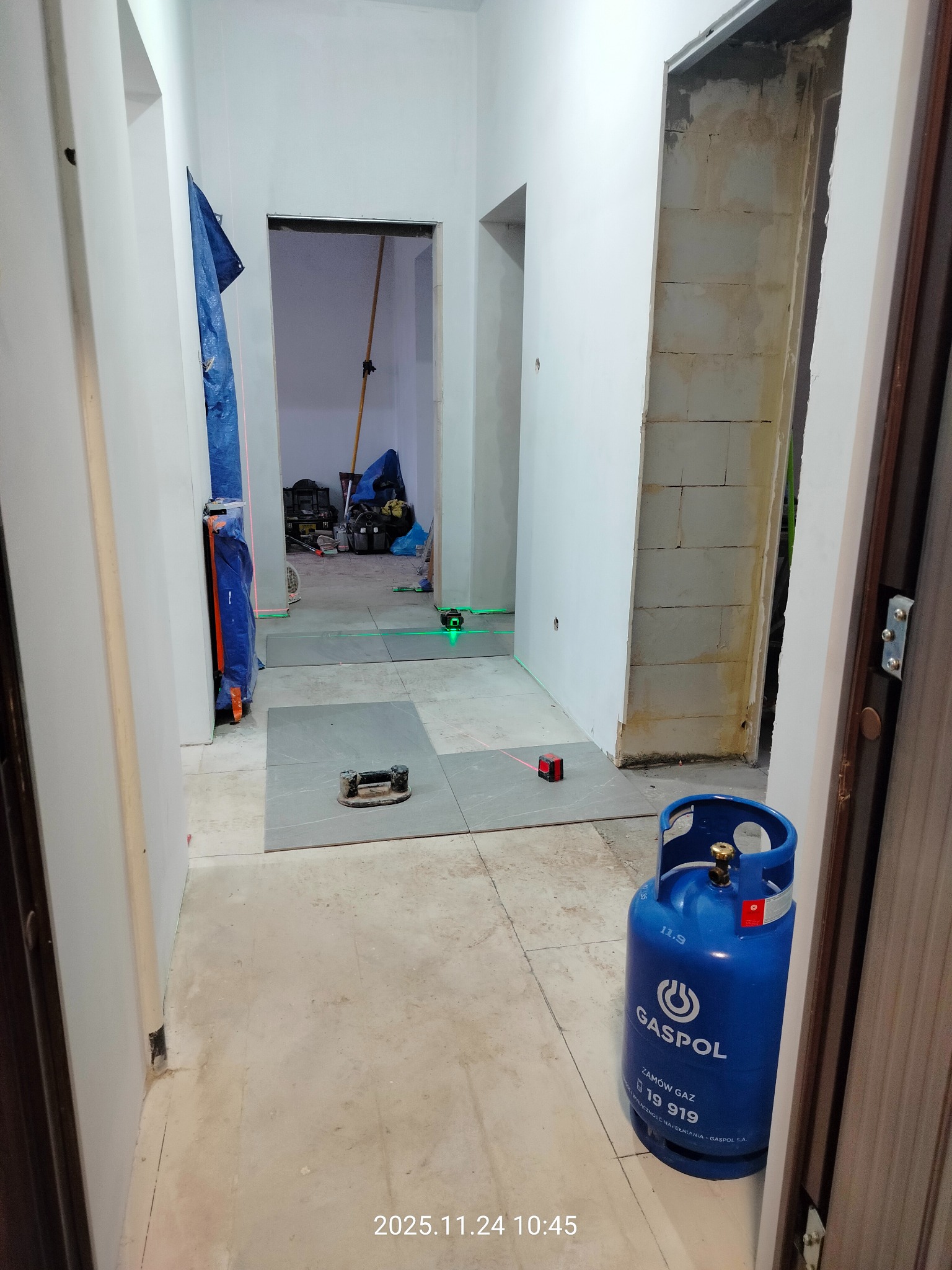 Korytarz w trakcie układania szarych płytek podłogowych. Widoczny laser krzyżowy, butla gazowa i narzędzia. Otwór drzwiowy w stanie surowym.