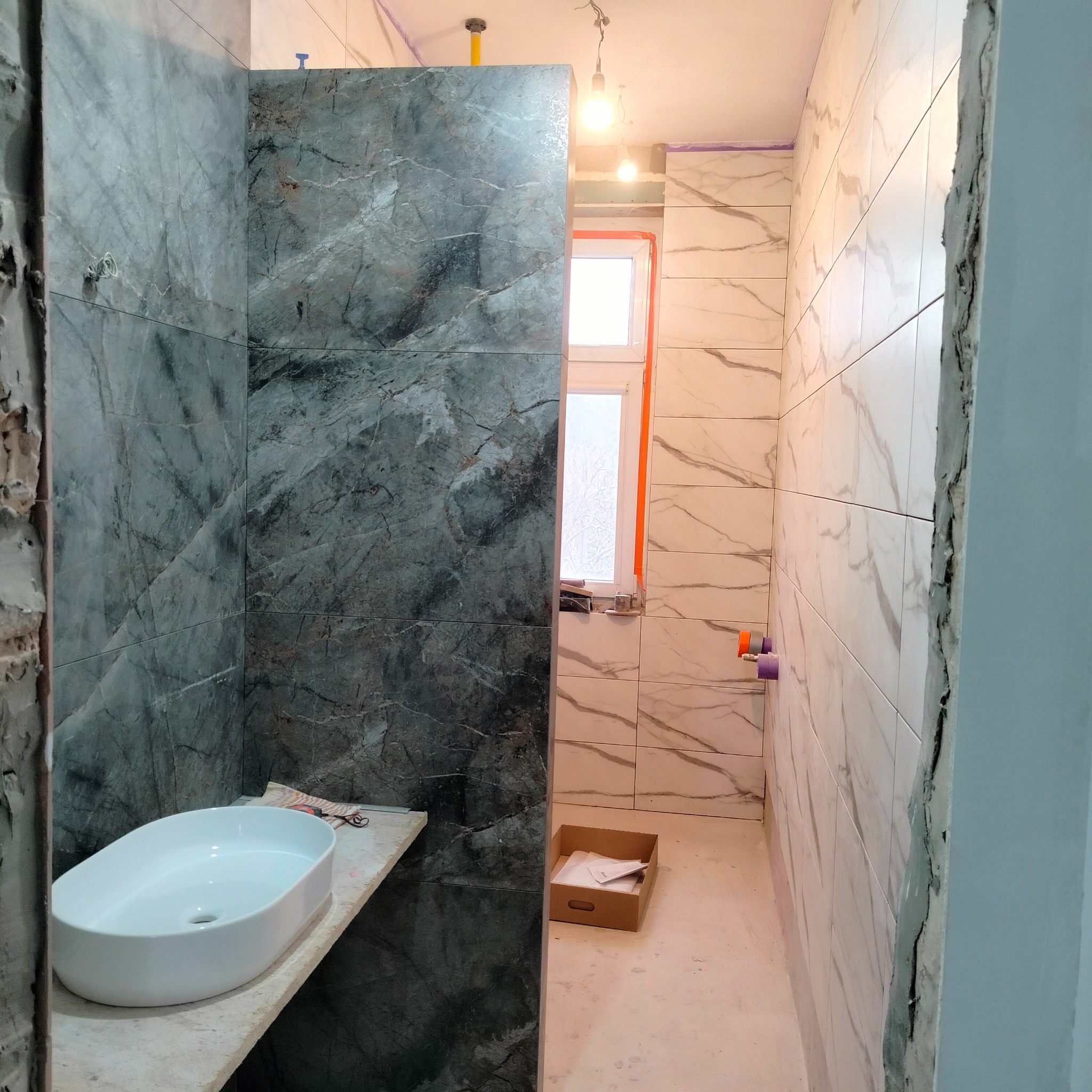 Łazienka w trakcie wykończenia: marmurowe płytki ścienne w odcieniach szarości i bieli, umywalka nablatowa, okno z pomarańczową ramą. Widoczne elementy instalacji.