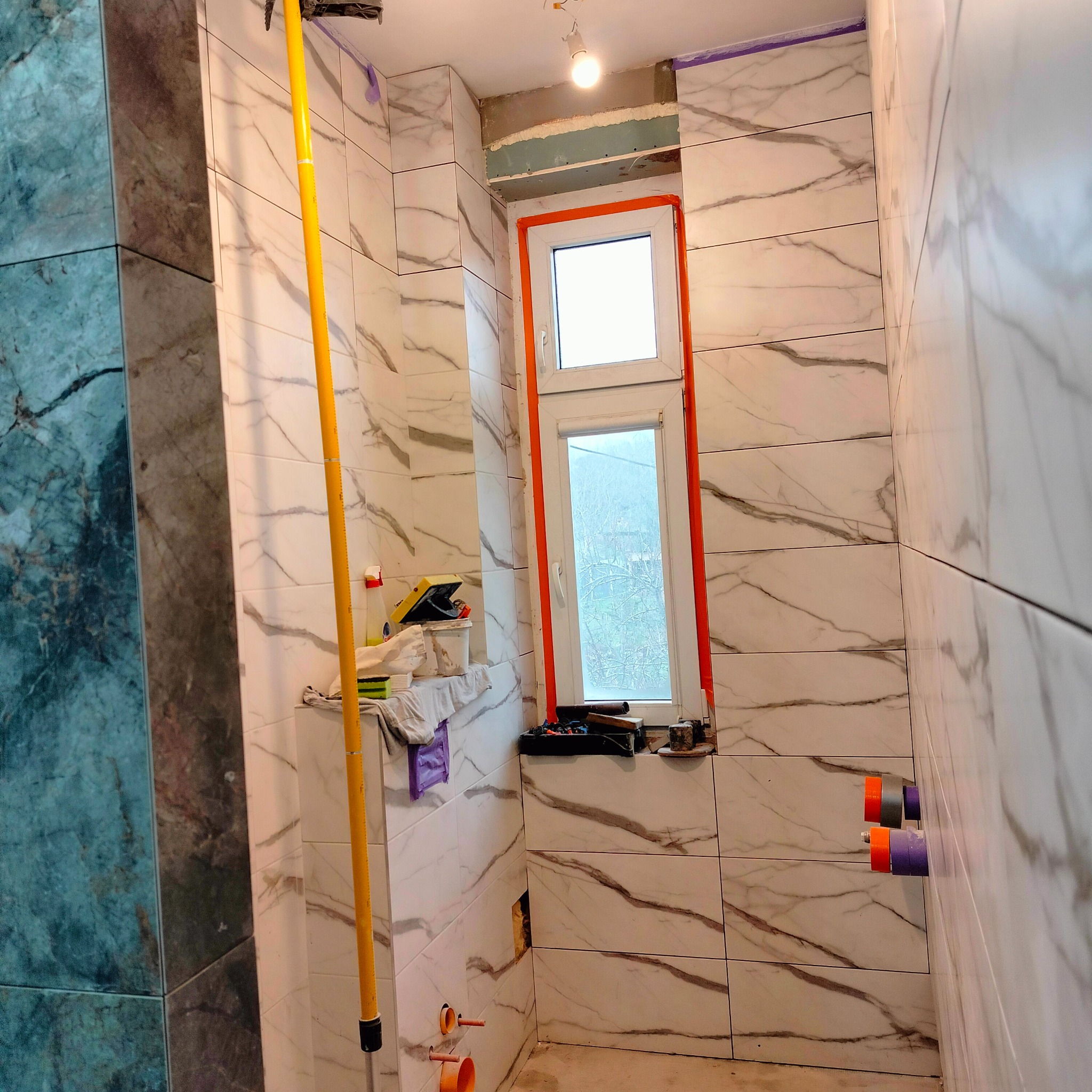 Łazienka w trakcie wykańczania: białe płytki imitujące marmur na ścianach, okno z pomarańczową ramą, widoczne narzędzia i akcesoria glazurnicze, rury w ścianie.