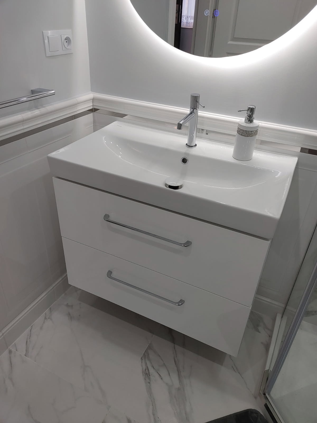 Biała umywalka z szafką i chromowaną armaturą w nowoczesnej łazience z marmurową podłogą i podświetlanym lustrem. Minimalistyczny design.